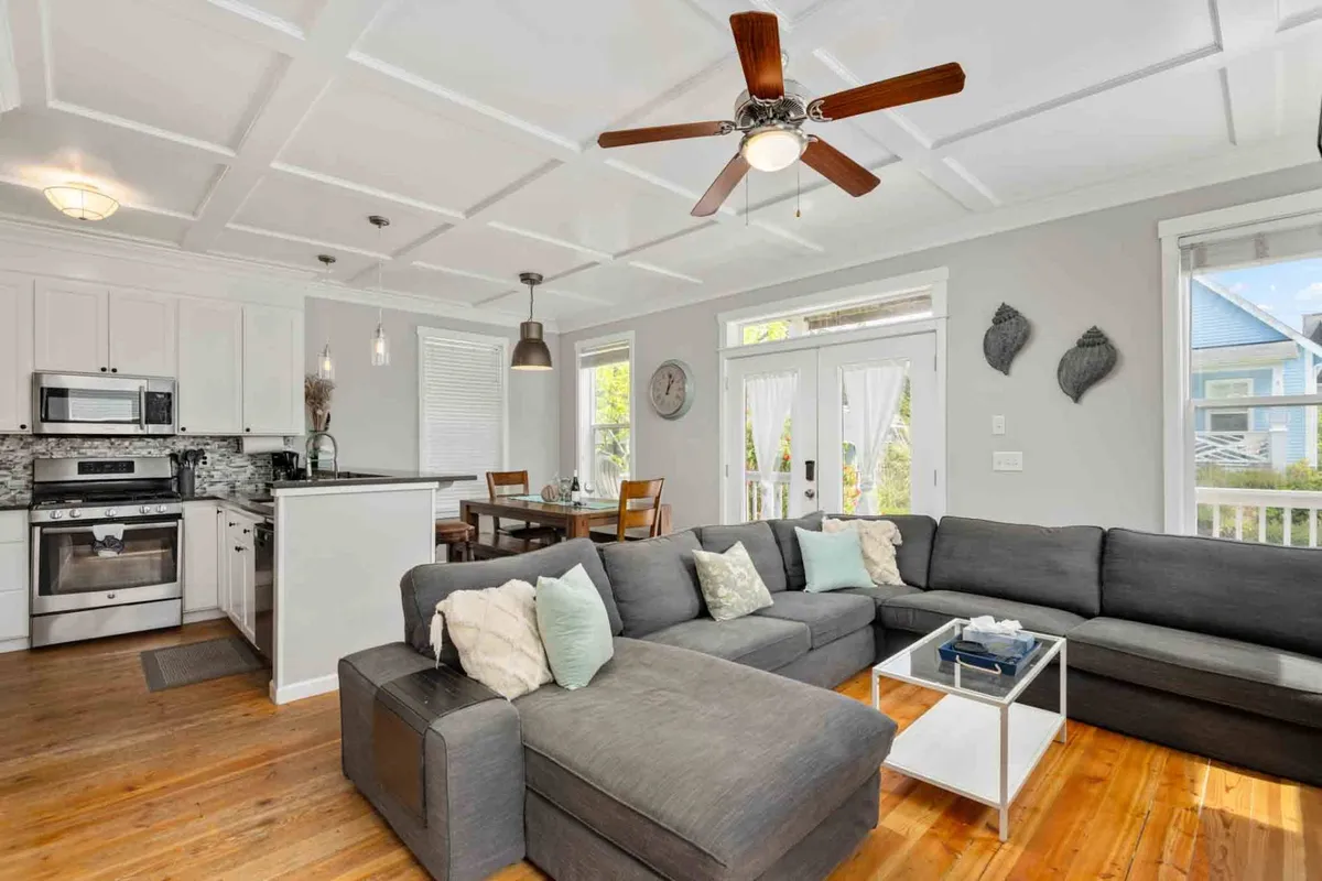 The image showcases a welcoming open-concept living space featuring a plush gray sectional sofa, a modern kitchen with stainless steel appliances, and a dining area flooded with natural light from large windows. The layout is designed for comfort and togetherness, perfect for family gatherings, cozy evenings, and memorable meals.