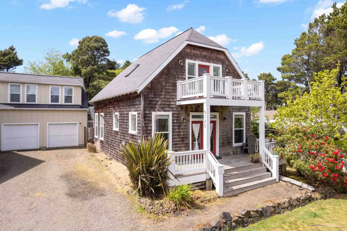 This exterior image captures the front porch of Bella Beach Retreat, showcasing welcoming wooden siding, white railings, and colorful flowers in bloom. The porch creates a connection to the outdoors and hints at areas where guests—and their pets—might relax or play together.