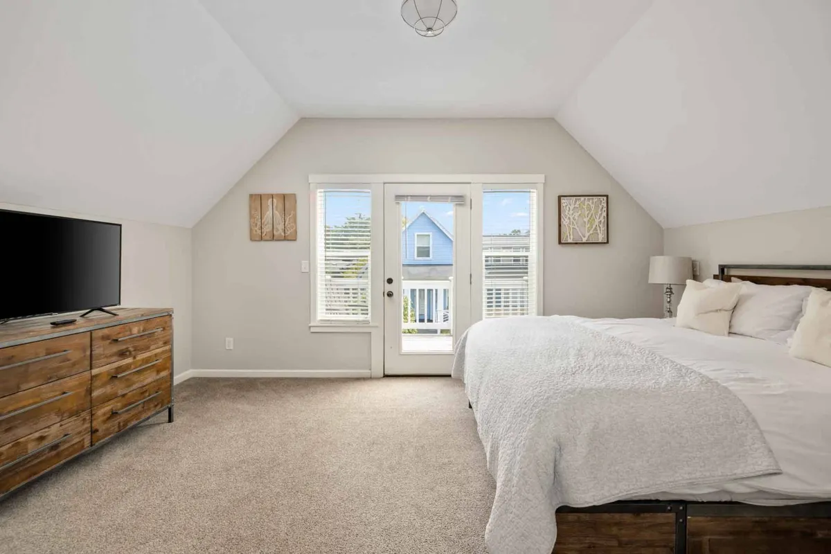 This image features a cozy bedroom with a bed, television, and a door leading out to a balcony. The presence of natural light streaming through the glass door hints at easy access to outdoor spaces and an airy, inviting atmosphere. The setup suggests a seamless connection between interior relaxation and enjoying the outdoors, such as stepping out for fresh air or a morning coffee while taking in nature.