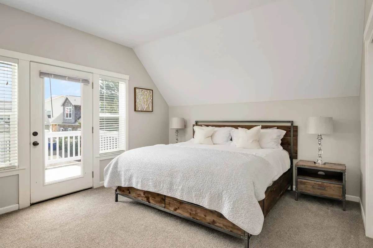 This image displays a serene king-sized bed dressed in white bedding, flanked by nightstands and lamps, with large windows that flood the room with natural light. The space exudes tranquility and provides a sense of privacy, ideal for a primary suite retreat.
