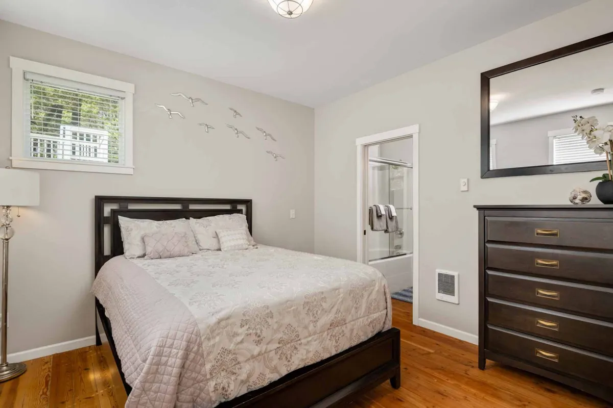 Featuring a cozy bed with decorative pillows, a framed mirror, and bird-themed wall art, this bedroom radiates comfort and quiet charm. Natural light streams in, creating an inviting space for relaxation and rejuvenation.
