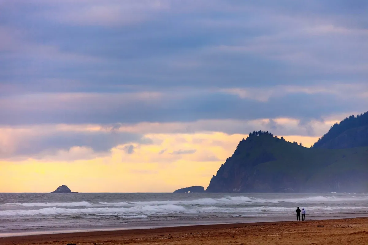 A picturesque coastal view featuring gentle ocean waves, distant cliffs, and two figures strolling along the beach at sunset. The scene is open and expansive, capturing the romantic and adventurous spirit of exploring the Oregon Coast.