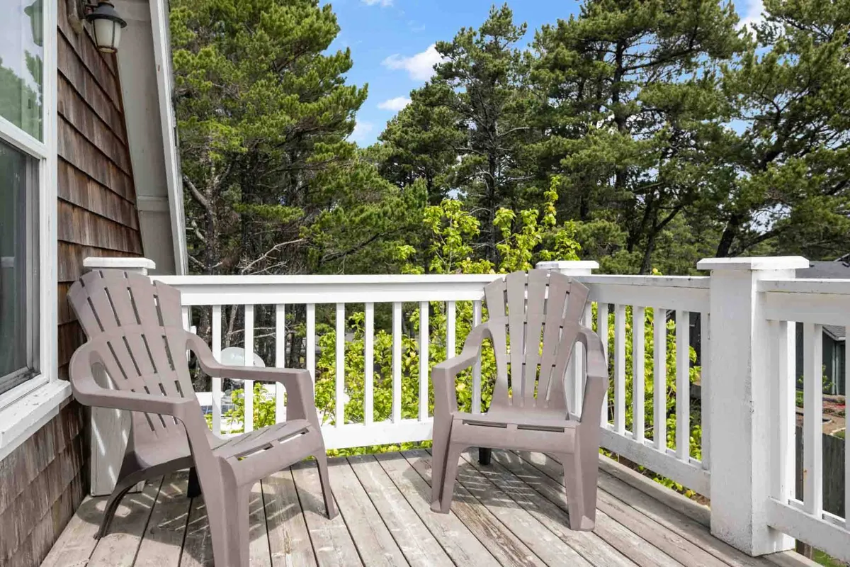 A peaceful exterior view of a wooden deck nestled among trees, outfitted with two Adirondack chairs. The natural setting is serene and inviting, ideal for quiet mornings or sunset conversations surrounded by coastal nature.
