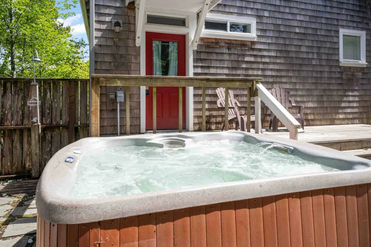 A tranquil exterior scene featuring a wooden deck with a bubbling hot tub as the centerpiece. The inviting red door and lush greenery surrounding the area create a private, rejuvenating atmosphere ideal for outdoor relaxation. Comfortable seating on the deck hints at alfresco dining and gatherings.