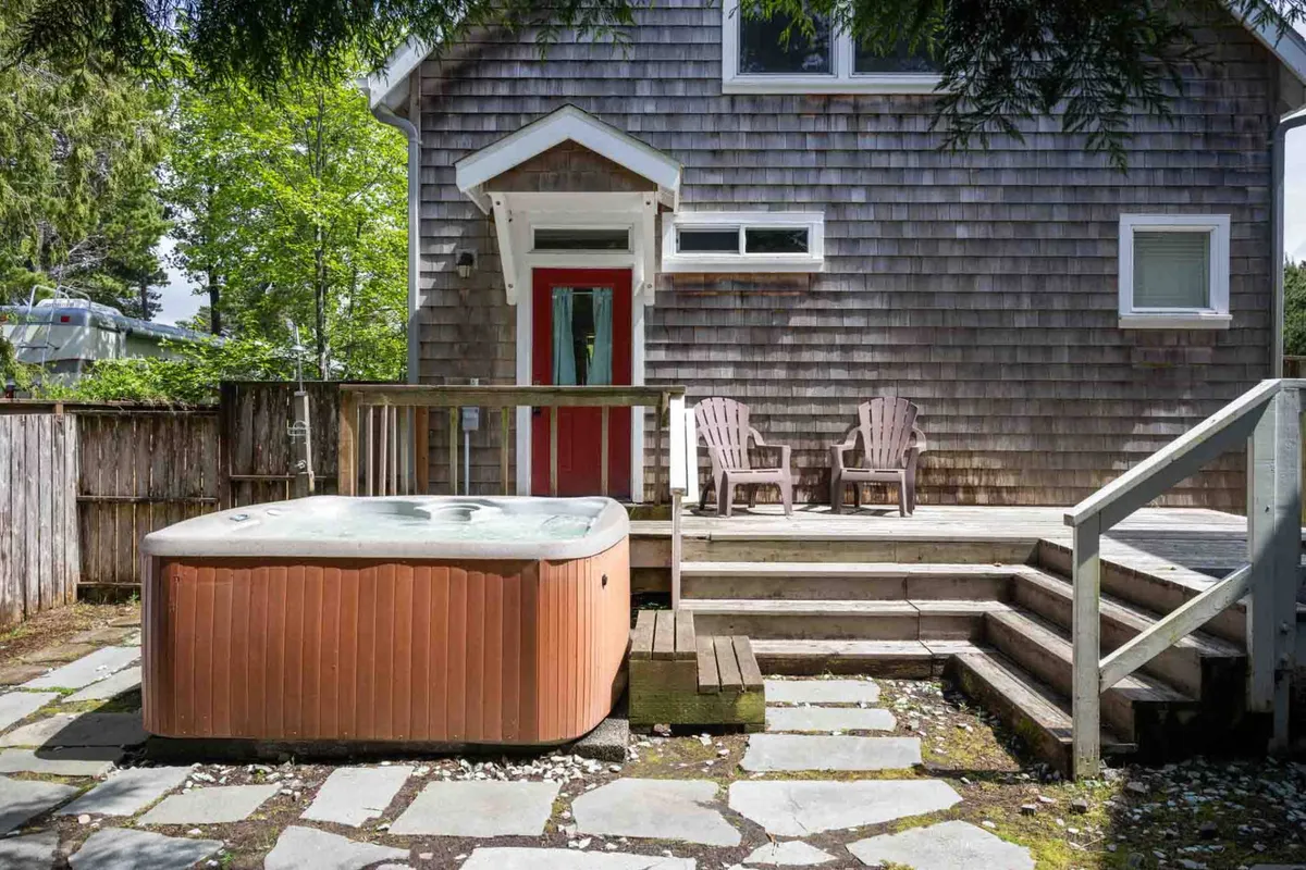A welcoming wooden deck featuring a bubbling hot tub, Adirondack chairs, and a backdrop of tall trees, with a distinctive red door lending charm. The setting conveys both relaxation and community gathering possibilities.